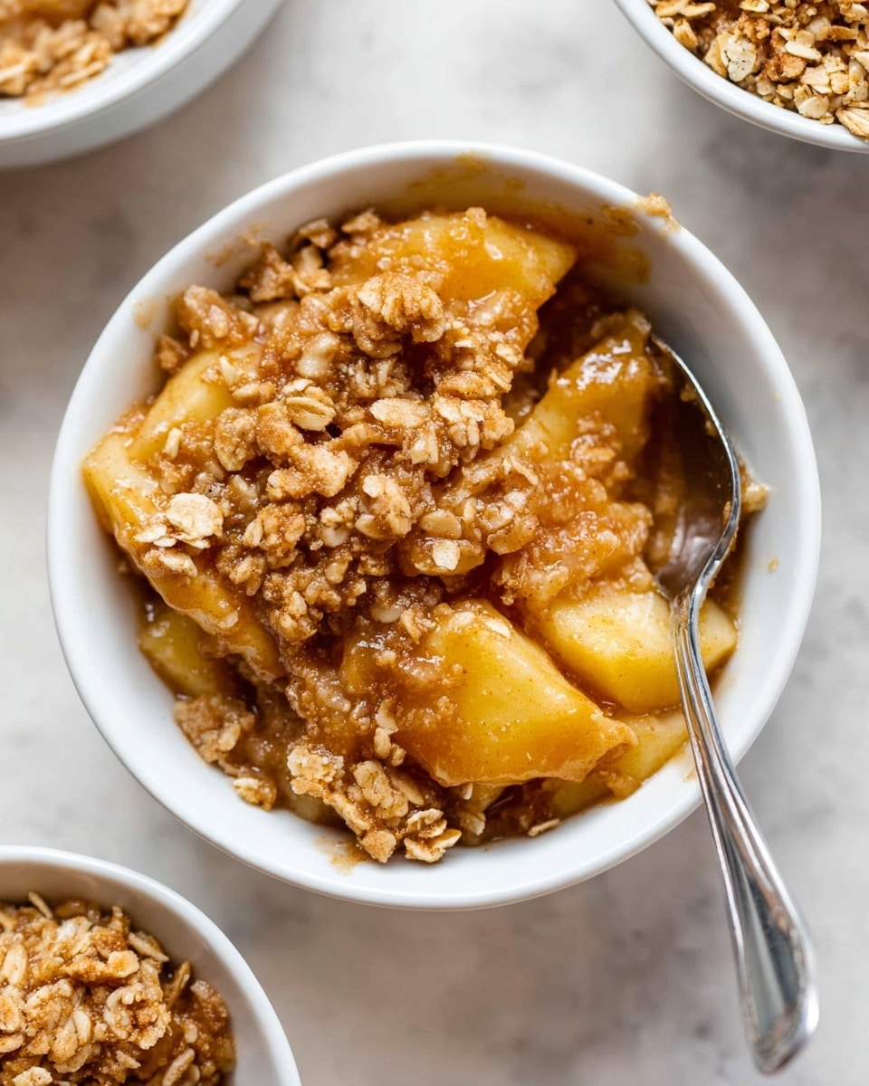 A white bowl holds a serving of apple crisp with two clear layers. The bottom layer is made of juicy, golden apple chunks covered in a thick, syrupy glaze. On top, there is a crumbly, golden-brown oat topping with visible rolled oats and a crunchy texture. A silver spoon rests inside the bowl, partially submerged in the apple and oat mixture. The bowl is placed on a white marbled surface with blurred parts of other bowls around it. Photo taken with an iphone --ar 4:5 --v 7