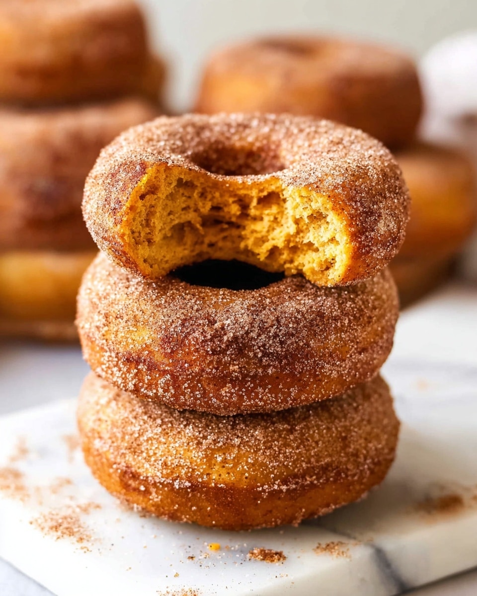 A stack of three round doughnuts with a light brown cinnamon sugar coating is placed on a white marbled surface. The top doughnut has a large bite taken out, showing a soft, moist, orange-yellow interior crumb. The doughnuts have a rough texture from the sugar and cinnamon on the surface. In the blurred background, more doughnuts are visible. photo taken with an iphone --ar 4:5 --v 7