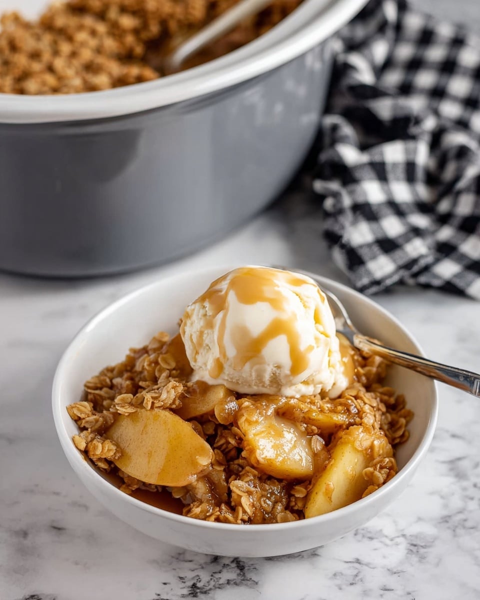 A white bowl filled with warm apple crisp, showing soft, golden-brown apple slices mixed with a thick layer of oat crumble on top, which has a sticky caramel color and textured oat pieces. Sitting on the oat crumble is a scoop of melting white vanilla ice cream covered lightly with creamy beige caramel sauce. A silver spoon rests on the edge of the bowl. In the background, a gray crockpot with a white interior is filled with more apple crisp, and a black and white checkered cloth is partly visible on a white marbled surface. Photo taken with an iphone --ar 4:5 --v 7