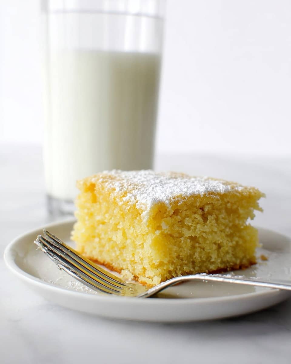 A single square piece of yellow, soft, and moist cake with a slightly crumbly texture is placed in the center of a white plate. The cake has a light dusting of powdered sugar on top, adding a delicate white layer. A silver fork rests on the edge of the plate, with its handle pointing toward the bottom left. In the background, slightly blurred, is a tall glass filled with white milk. The scene is set on a white marbled surface, creating a clean and simple look. Photo taken with an iphone --ar 4:5 --v 7