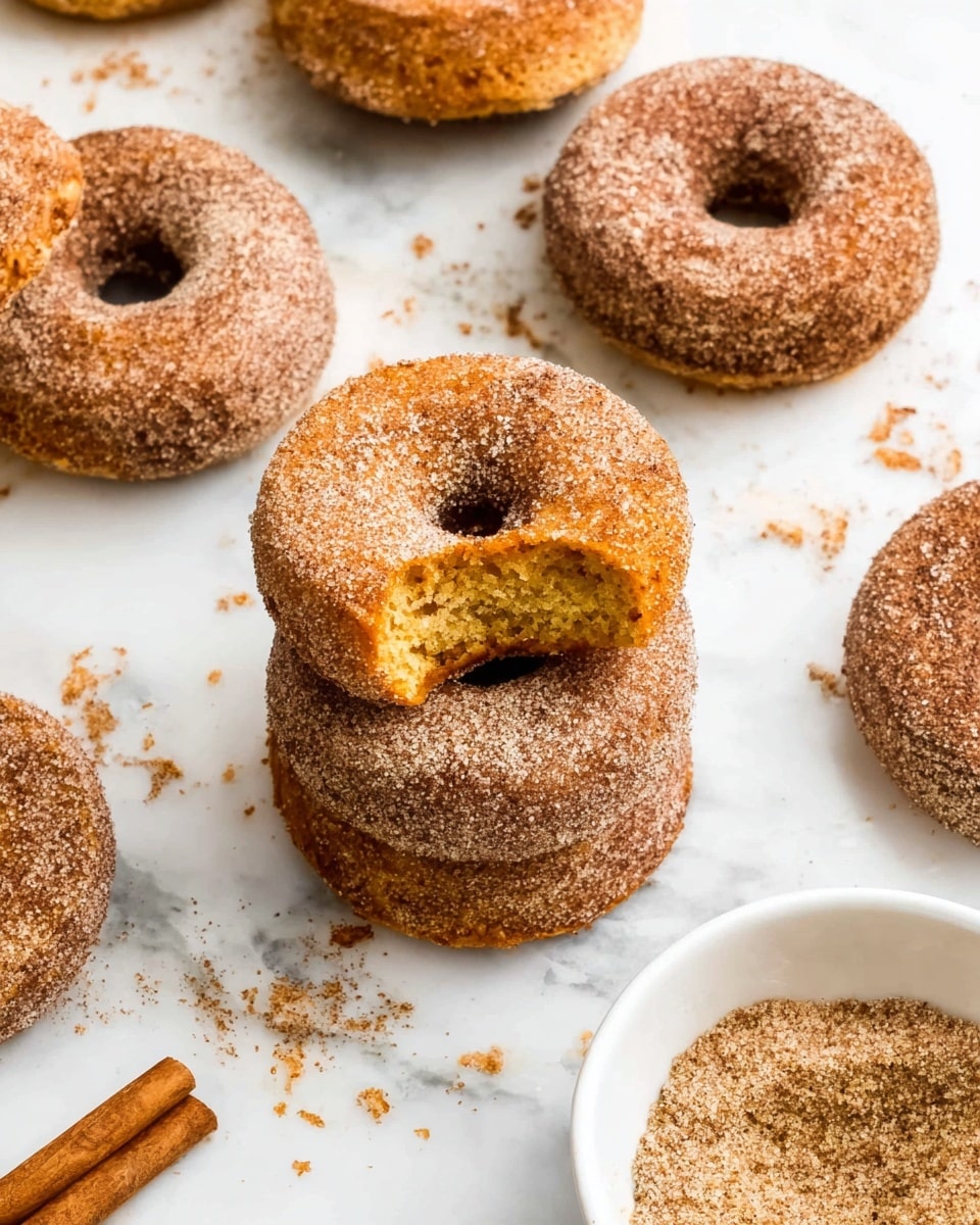 The image shows several cinnamon sugar-coated donuts arranged on a white marbled surface. Each donut has a rough, textured light brown outer layer covered in cinnamon sugar. One donut is placed on top of another at the center, with the top donut showing a bite taken out, revealing a soft, yellow, cake-like inside. Around the donuts, there are some crumbs scattered, and a white bowl on the right side filled with more cinnamon sugar. A few cinnamon sticks are placed at the bottom left corner of the image. Photo taken with an iphone --ar 4:5 --v 7