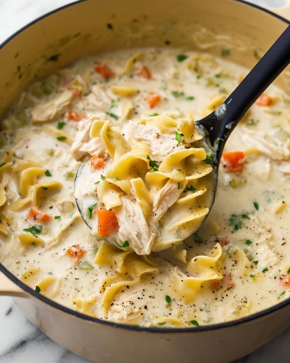 The image shows a close-up of a creamy chicken noodle soup in a large round pot with a light beige inside. The soup has a thick white cream base mixed with tender pieces of white chicken meat, short wide yellow egg noodles, small orange carrot cubes, and green parsley bits scattered throughout. A black ladle lifts a scoop full of the soup, showing the mix of smooth creamy sauce, noodles curled and layered, and fresh diced vegetables. The soup looks warm and hearty, with some black pepper seasoning visible on top. The background is a white marbled texture. Photo taken with an iphone --ar 4:5 --v 7