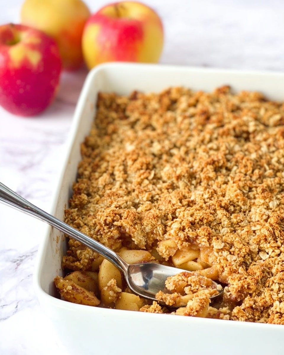 A white rectangular baking dish filled with a golden brown apple crumble that has a textured oat and cinnamon crumb topping covering a layer of soft, light brown baked apple chunks underneath; a shiny silver spoon rests inside the dish on the left side, and three red and yellow apples sit slightly blurred in the white marbled background behind the dish. photo taken with an iphone --ar 4:5 --v 7