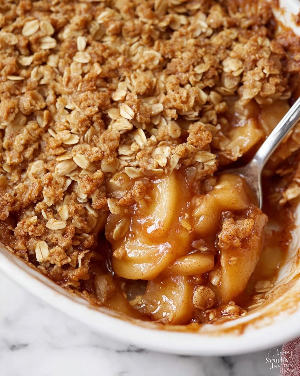 The image shows a close-up of a baked apple crisp in a white dish, revealing two main layers. The bottom layer is made of juicy, soft apple slices coated in a glossy, cinnamon-spiced syrup with a light brownish color. The top layer is a thick, crunchy oat crumble with golden brown clusters and small rolled oats scattered throughout. The edges of the dish show some syrup spilling slightly over the apples, adding a sticky texture. The spoon in the dish scoops some of the apple layer and crumble, emphasizing the contrast between the soft fruit and crisp topping. The background features a white marbled texture. photo taken with an iphone --ar 4:5 --v 7