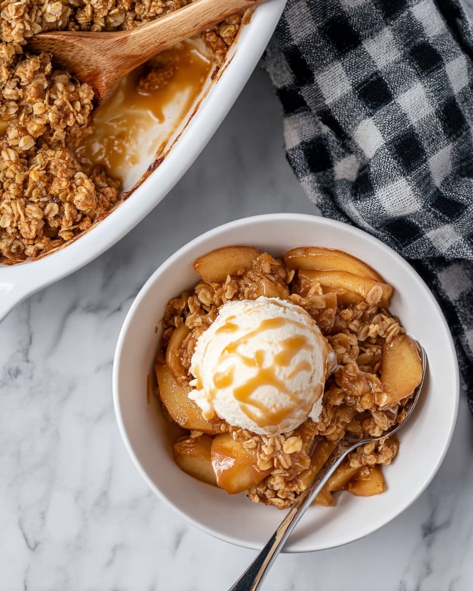 A white bowl holds a warm apple crisp dessert with two main visible layers: a bottom layer of soft, sliced apples in a light brown syrup and a top layer of golden, crumbly oat topping with a slightly crunchy texture. Sitting on top of the apple crisp is a round scoop of creamy vanilla ice cream with a smooth texture, slightly melting and drizzled with a light caramel sauce. The bowl is on a white marbled surface next to a silver spoon resting inside. A white baking dish filled with more apple crisp is partially visible in the upper left corner, with a wooden spoon resting inside. A black and white checkered cloth is placed beside the bowl. photo taken with an iphone --ar 4:5 --v 7