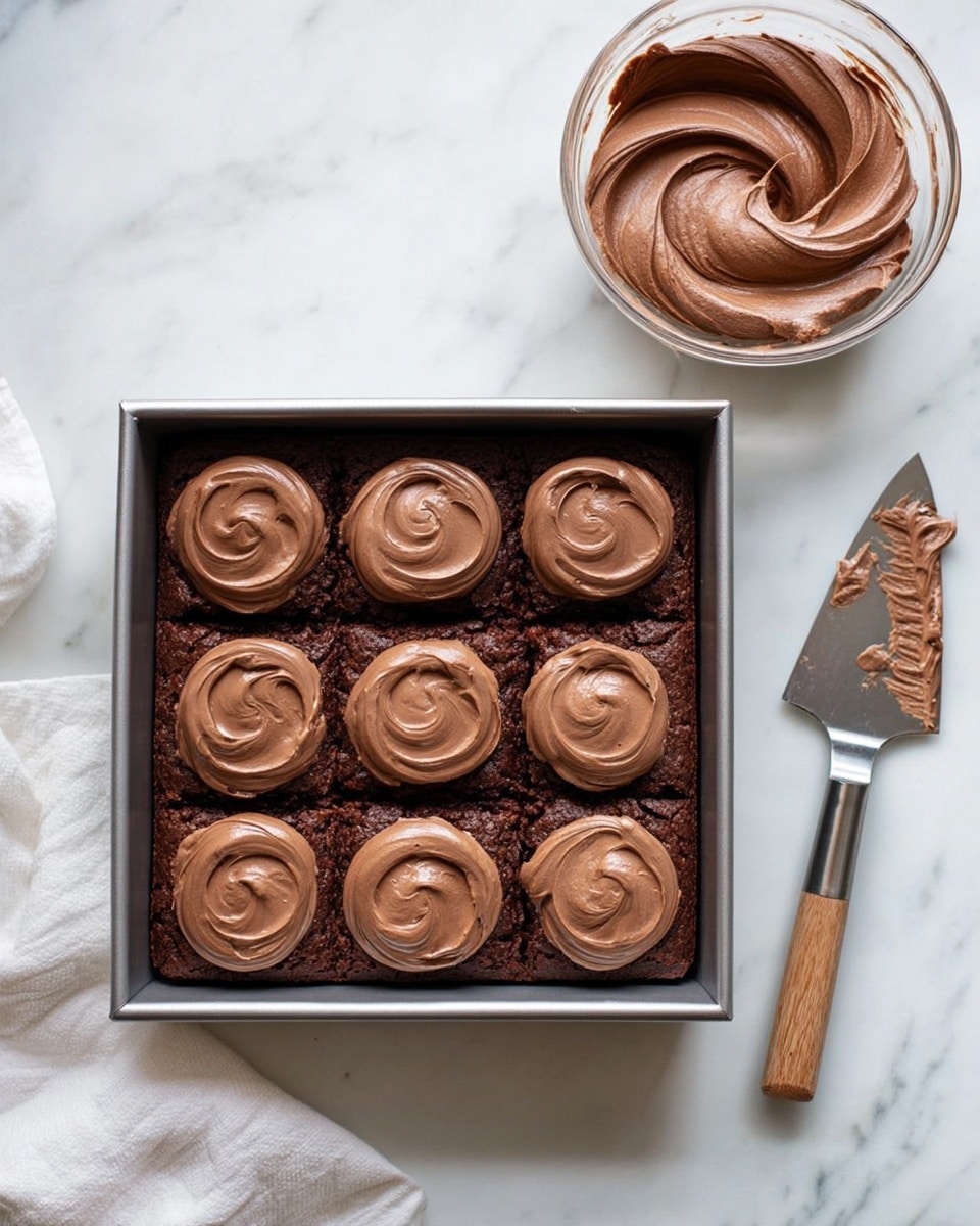 A square dark brown chocolate brownie sits in a metal baking pan on a white marbled surface, topped with nine dollops of smooth, light chocolate frosting arranged in a loose grid. Each dollop is circular with a slightly swirled texture and raised edges, sitting evenly over the brownie base. To the top right of the pan is a clear glass bowl filled with the same light chocolate frosting, showing a creamy and swirled texture. To the right of the pan lies a metal spatula with a wooden handle, with traces of chocolate frosting on the blade. A white cloth is partially visible to the bottom left corner. photo taken with an iphone --ar 4:5 --v 7