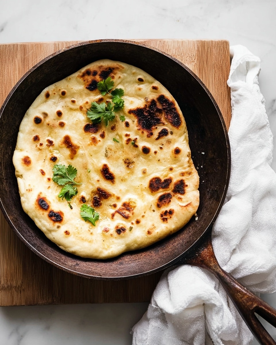 A single round flatbread with a golden-brown, lightly charred surface sits inside a dark, rustic frying pan. The flatbread has a soft, uneven texture with some puffed areas and darker burnt spots scattered on top. On one side, a few fresh cilantro leaves add a touch of green. The frying pan rests on a light wooden board, placed on a white marbled surface. A crumpled white cloth is visible beside the handle of the pan. photo taken with an iphone --ar 4:5 --v 7