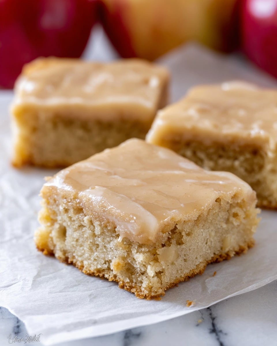 The image shows three square pieces of a light brown cake with a slightly shiny glaze on top. Each piece has two visible layers: a soft, crumbly cake base in a pale beige color and a thin layer of light brown frosting or glaze on top. The cake layers look moist and slightly dense with a few small chunks visible inside. The pieces are placed on white parchment paper, resting on a white marbled surface, and there are blurred red apples in the background. Photo taken with an iphone --ar 4:5 --v 7