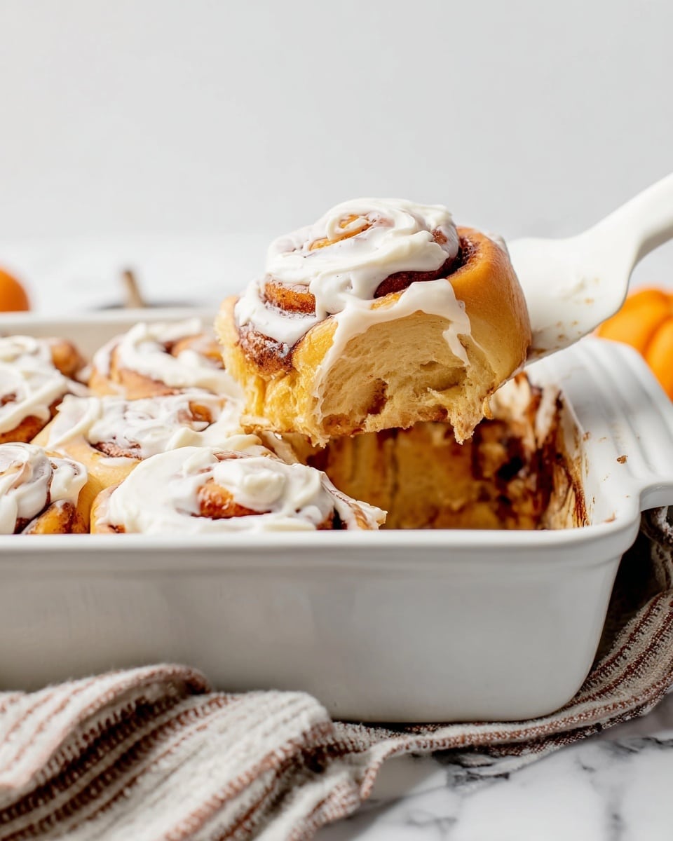 The image shows a white rectangular baking dish filled with a batch of soft cinnamon rolls. The cinnamon rolls have a golden-brown color with a visible spiral pattern on top and are generously covered with thick white cream cheese frosting. One cinnamon roll is being lifted out with a white spatula, showing its fluffy, moist interior with a bit of frosting on top. The dish is set on a white marbled surface, and a textured cloth with stripes is partly visible beneath the dish. Photo taken with an iphone --ar 4:5 --v 7