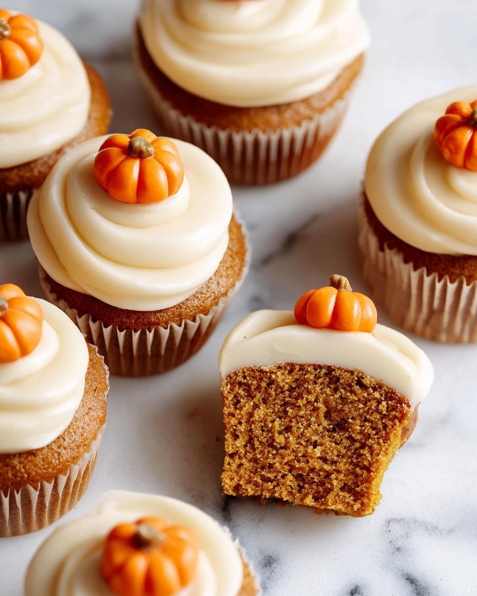 The image shows several pumpkin cupcakes with two layers: a moist, spongy orange-brown cake base and a thick, smooth, swirled white frosting on top. Each cupcake is topped with a small, shiny orange pumpkin-shaped candy placed near the edge of the frosting. One cupcake is cut in half to show the cake texture beneath the frosting. The cupcakes are arranged on a white marbled surface, with additional small pumpkin candies scattered around. Photo taken with an iphone --ar 4:5 --v 7