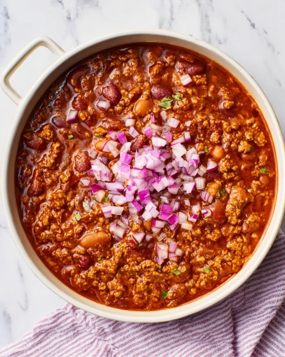 The image shows a white deep round dish filled with a thick, textured chili. The chili has a rich, reddish-brown sauce with visible ground meat and beans. There is a topping of finely chopped red onions scattered evenly on top, adding a pop of pale purple and white color. The dish is placed on a white marbled surface with a striped cloth partially visible beside it. The lighting highlights the glossy and hearty texture of the chili, making it look warm and ready to eat. Photo taken with an iphone --ar 4:5 --v 7
