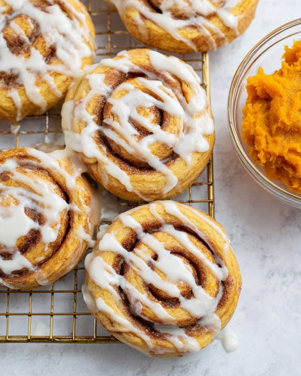 The image shows several round cinnamon rolls arranged on a gold cooling rack over a white marbled surface. Each roll has a soft, golden brown dough base with visible cinnamon filling spiraled inside. On top, there is a glossy white icing drizzled unevenly, creating thin and thick stripes that follow the spiral pattern of the roll. To the right side, a clear glass bowl filled with bright orange pumpkin puree is partially visible. The overall look is warm and inviting with a fresh-baked texture. photo taken with an iphone --ar 4:5 --v 7