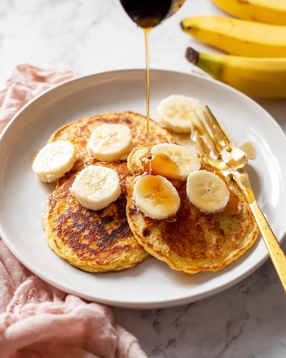 A white plate on a white marbled surface holds three golden brown pancakes arranged slightly overlapping. The pancakes have a soft, slightly uneven texture with some darker browned spots. On top and around the pancakes are several thick banana slices, pale yellow with soft edges. A thin stream of amber syrup is shown pouring over the middle pancake, creating a shiny, sticky layer that spreads outward. A gold fork rests on the right side of the plate with its handle extending beyond the plate’s edge. In the background, there are a pair of yellow bananas and a light pink cloth partially visible. photo taken with an iphone --ar 4:5 --v 7