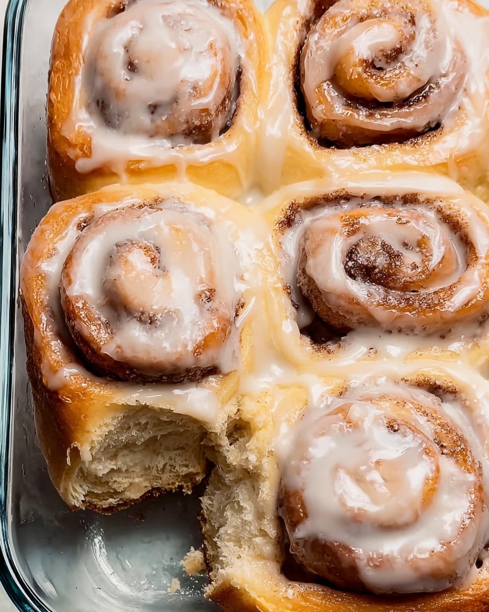 The image shows four soft, thick cinnamon rolls in a glass baking dish with a white marbled background. Each roll is golden brown on the outside, with light brown cinnamon swirls visible on top. They are generously coated with smooth, shiny white icing that drips slightly down the sides. One roll in the bottom left corner has a piece taken out, revealing a fluffy, light beige interior with a tender texture. The cinnamon rolls are close together, filling the dish evenly. Photo taken with an iphone --ar 4:5 --v 7