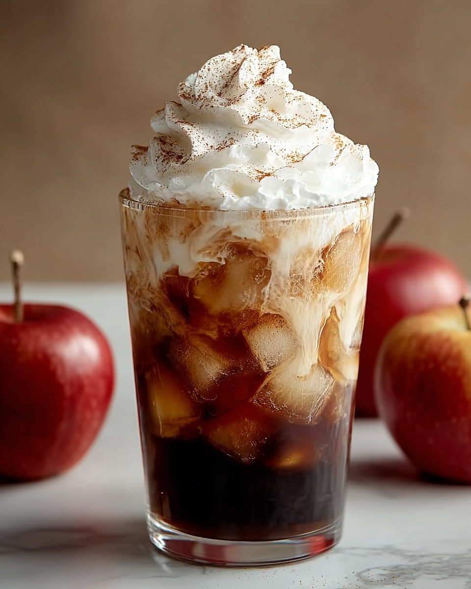 A clear glass filled with a dark brown liquid at the bottom, followed by a layer of melting ice cubes creating a mix of light and dark brown swirls, topped with a large, fluffy swirl of white whipped cream sprinkled lightly with brown cinnamon powder; the glass is set on a white marbled surface with two red apples with brown stems placed blurred in the background on either side. photo taken with an iphone --ar 4:5 --v 7