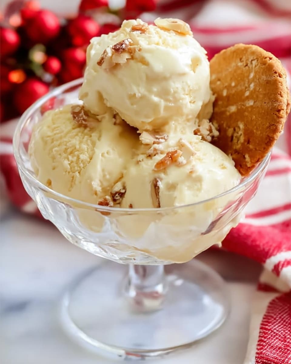 A clear glass dessert cup holds three scoops of creamy light yellow ice cream mixed with small pieces of nuts, giving a slight grainy texture to the smooth surface. A round cookie with nut bits is placed leaning inside the cup on the right side. The cup sits on a white marbled surface with a soft focus red and white striped cloth visible underneath and around it. The overall look is bright and inviting with soft natural light. Photo taken with an iphone --ar 4:5 --v 7