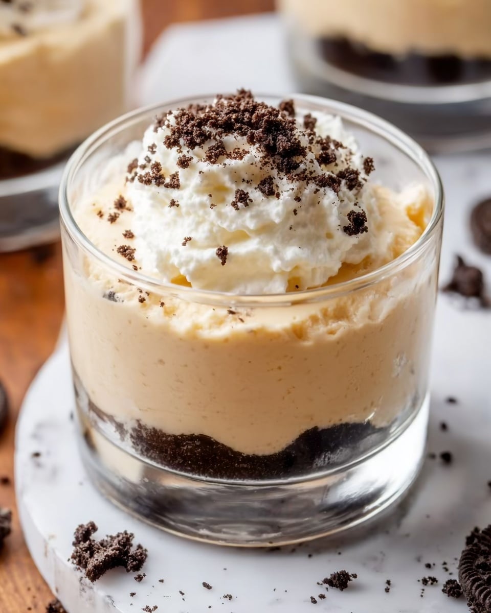 The image shows a clear glass with three layers of dessert standing on a white marbled surface. The bottom layer is a thick creamy light beige mousse with a smooth and slightly swirled texture. The middle layer is very similar in color and texture but appears a bit denser and slightly more whipped, filling most of the glass. On top, there is a generous white whipped cream swirl with soft, smooth peaks, sprinkled lightly with small golden crumbs. In the blurred background, two more similar glasses are visible, and next to the main glass, a small portion of a gold spoon rests on the surface. Photo taken with an iphone --ar 4:5 --v 7