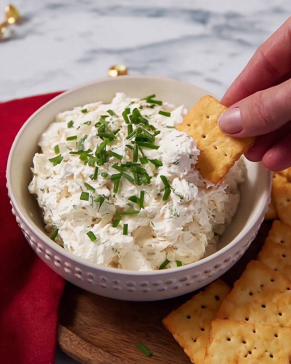 A round white bowl with small raised dots around the rim holds a thick, creamy white mixture with a slightly rough texture, topped with fresh green chopped chives. In the foreground, a woman's hand is dipping a small, round golden cracker into the creamy mixture. To the right, a wooden tray is partially visible, holding two square golden crackers with small holes and browned spots. The bowl and tray are set on a surface with a white marbled texture, and a red cloth is seen to the left. photo taken with an iphone --ar 4:5 --v 7