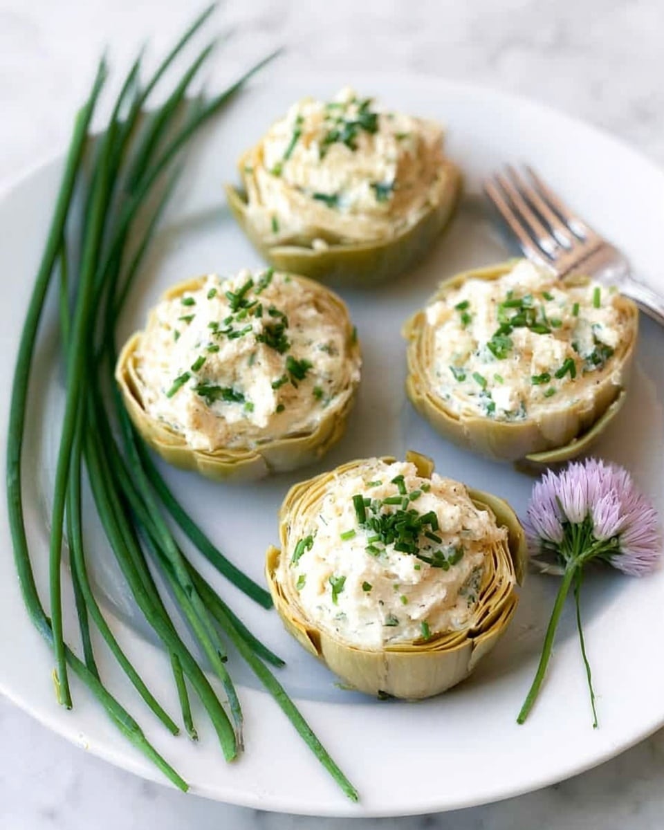 The image shows four stuffed artichoke halves placed on a white round plate. Each artichoke half is filled with a creamy white mixture that has a slightly lumpy texture, topped with small chopped green herbs sprinkled evenly. Next to the artichokes on the left side of the plate are several long green chive stalks and some chive flowers at the bottom. A silver fork rests on the upper right side of the plate. The background is a white marbled surface. photo taken with an iphone --ar 4:5 --v 7