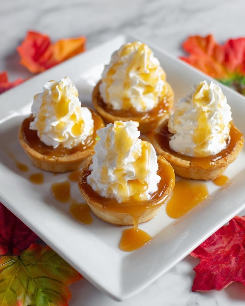 Four small round tartlets with a golden brown crust sit on a white square plate, each topped with a fluffy white layer of whipped cream. A shiny amber caramel sauce is drizzled over the whipped cream and pooling around the tartlets on the plate. The plate is placed on a white marbled surface scattered with colorful orange, red, and green autumn leaves. photo taken with an iphone --ar 4:5 --v 7
