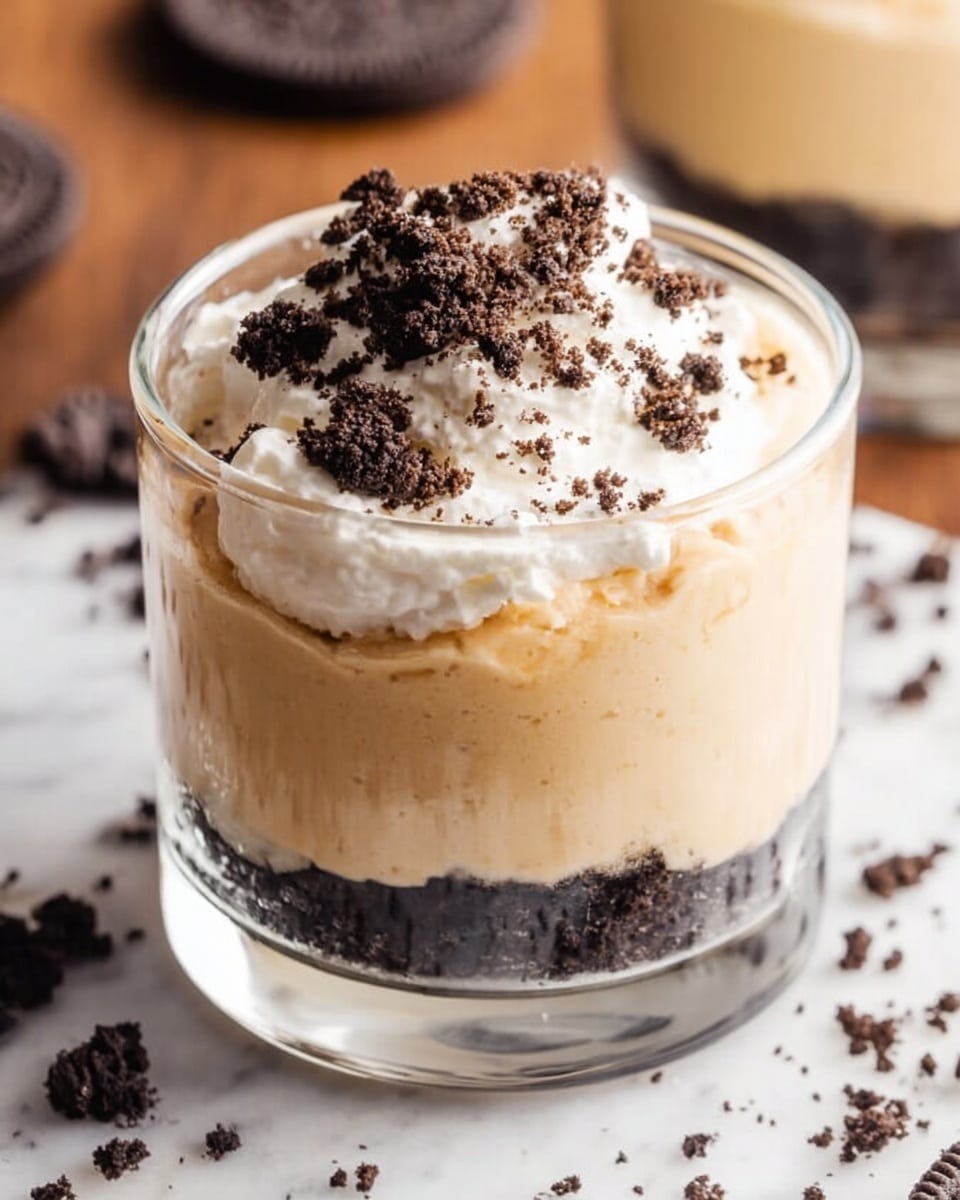 A clear glass cup filled with two main layers, the bottom layer is a thick, smooth light brown mousse with a creamy texture, topped with a generous swirl of bright white whipped cream that looks soft and fluffy, sprinkled with small golden crumbs. The cup sits on a white marbled surface with a shiny gold spoon nearby. In the background, there are blurred similar glass cups filled with the same dessert. photo taken with an iphone --ar 4:5 --v 7