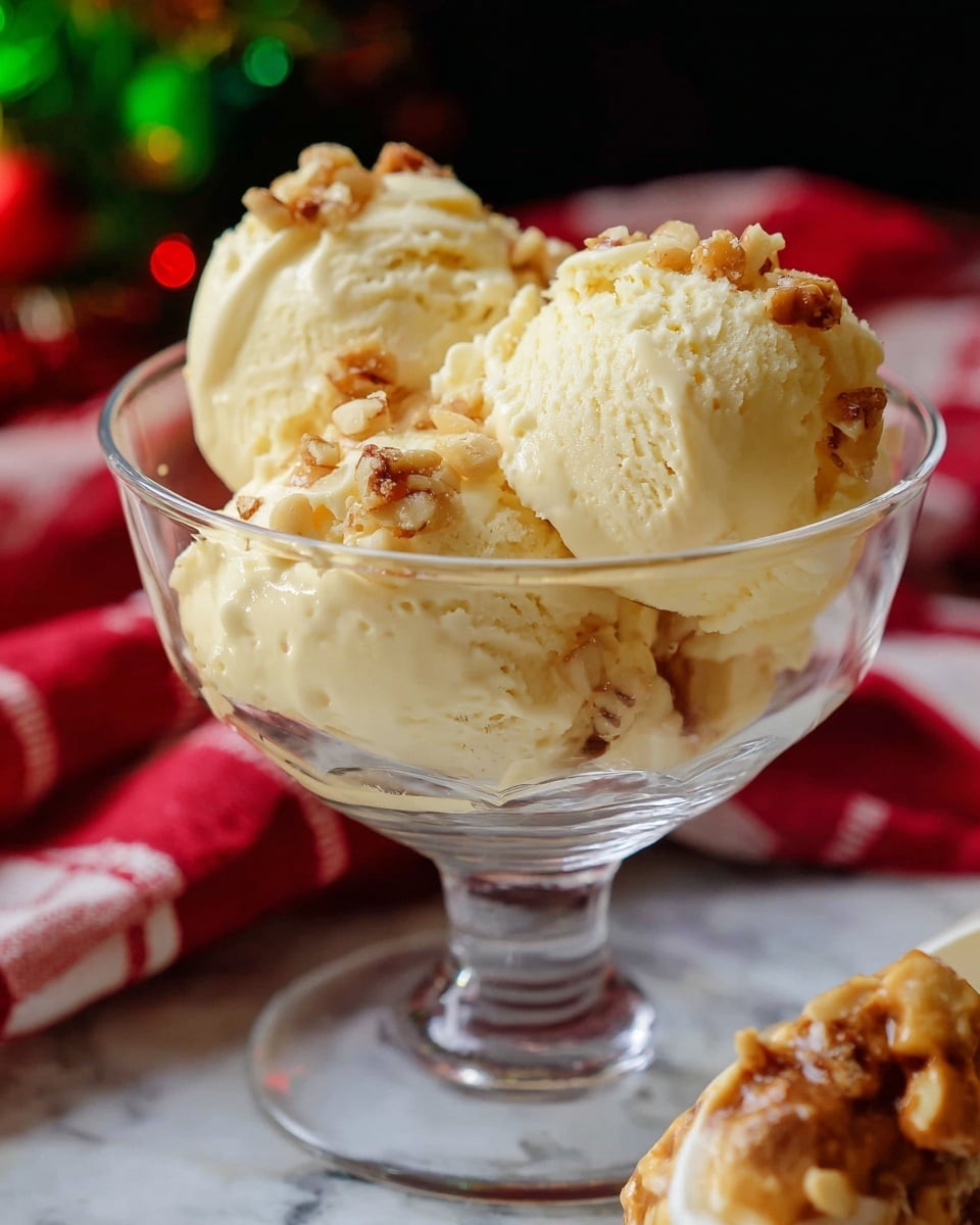 A clear glass dessert bowl with a short stem holds three scoops of pale yellow ice cream, each sprinkled with small crunchy pieces that look like nuts. At the back right side of the bowl, a scoop partially covered with caramel or toffee bits is visible. The glass rests on a white marbled surface with a red and white striped cloth nearby, and the background is dark with blurry hints of green and red colors. photo taken with an iphone --ar 4:5 --v 7