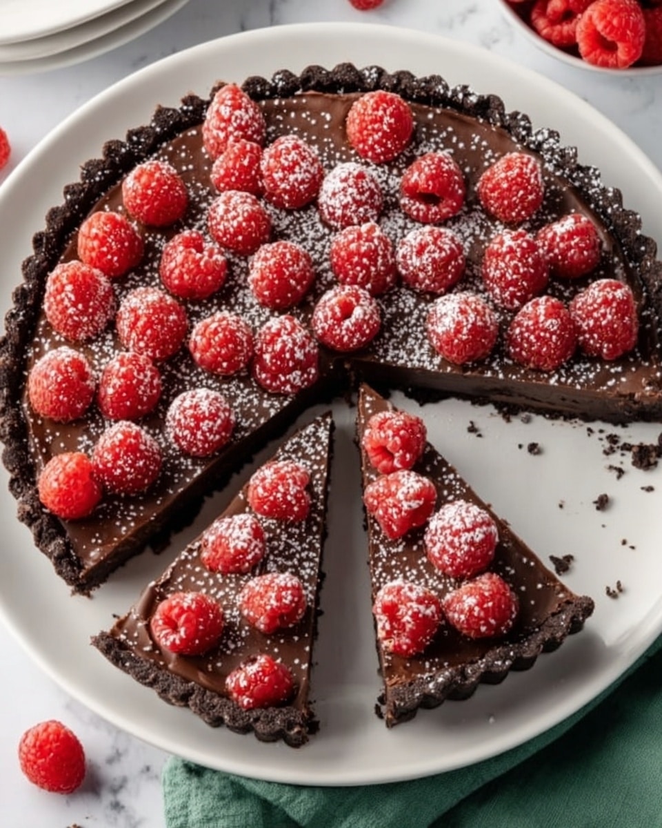 A close-up view of a chocolate tart with three distinct layers: the bottom layer is a dark crumbly crust, the middle layer is smooth, glossy dark chocolate filling, and the top layer is fresh, bright red raspberries evenly placed over the chocolate. Powdered sugar is being sprinkled over the tart, creating a light dusting on the raspberries and chocolate. Three loose raspberries sit on a white plate in the background alongside forks resting on a white marbled surface. Photo taken with an iphone --ar 4:5 --v 7