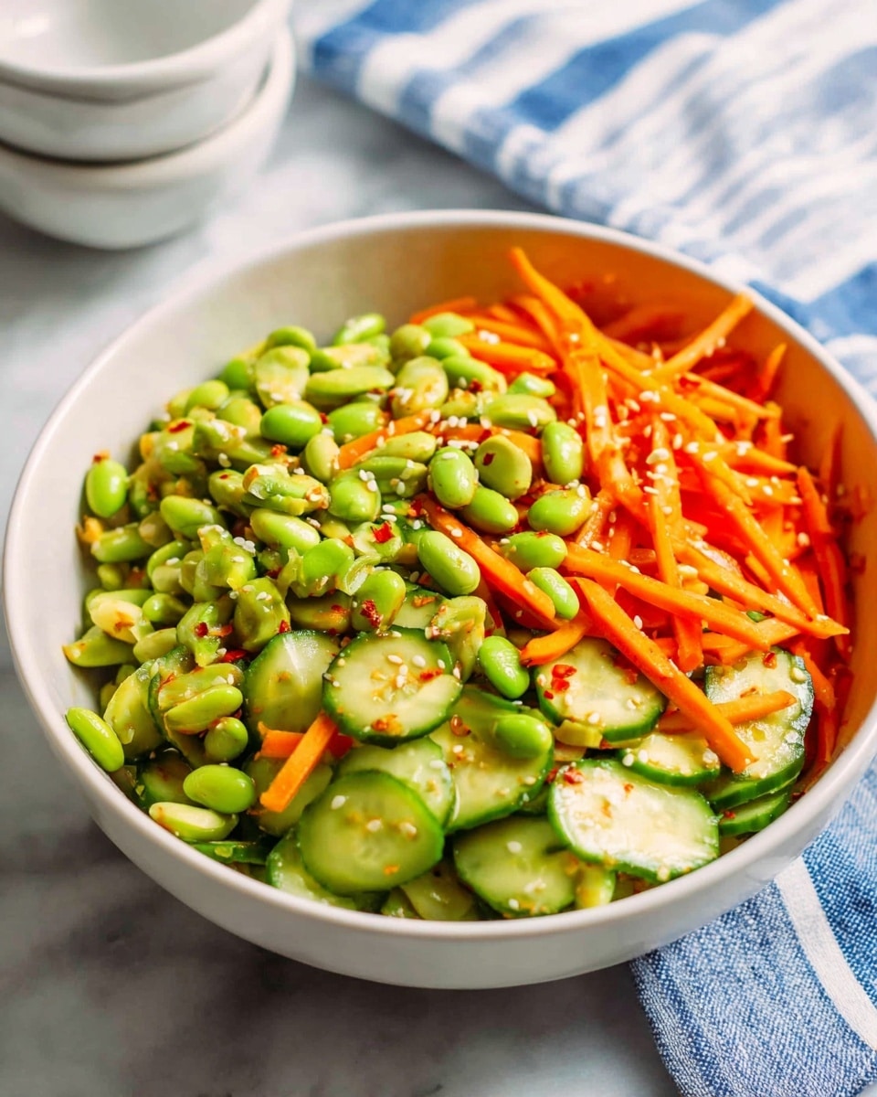 A white bowl filled with a colorful salad sits on a white marbled surface. The salad has three clear layers: the bottom layer is crunchy cucumber slices with light green and dark green edges, the middle layer includes bright orange thin carrot sticks, and the top layer shows many light green edamame beans scattered all over. Small white sesame seeds and tiny red chili flakes are sprinkled on the salad. In the background, blurred white cups are stacked and a towel with blue and white stripes is partly visible. photo taken with an iphone --ar 4:5 --v 7