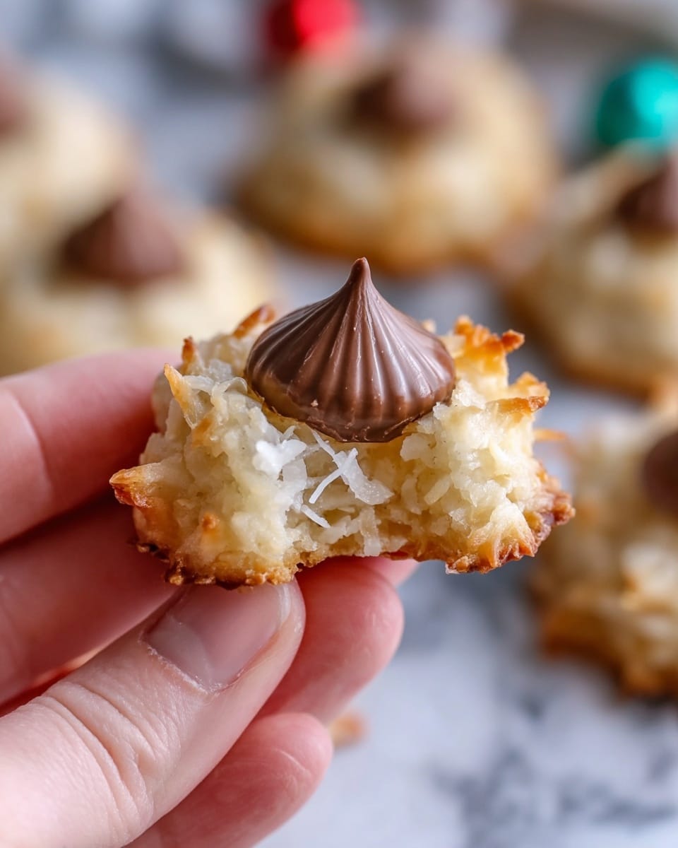 A close-up image of a woman's hand holding a bite-sized coconut cookie with a single milk chocolate kiss in the center. The cookie has a rough texture, golden brown edges with shredded white coconut visible, and a slightly crispy look. The chocolate kiss is smooth, dark brown, and placed on top of the cookie. The background shows other similar cookies slightly blurred on a white marbled surface. Photo taken with an iphone --ar 4:5 --v 7