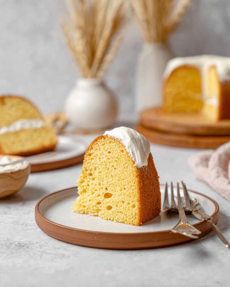 A close-up of a slice of yellow sponge cake with a smooth and creamy white filling in the center, standing upright on a round white plate with a brown rim and two silver forks beside it; the cake has a moist and airy texture with tiny holes throughout. In the blurred background, more slices of the same cake sit on similar plates, with wooden cutting boards and white vases holding dried wheat stalks placed on a white marbled surface. The scene is softly lit, emphasizing the golden color of the cake and the creamy white filling. photo taken with an iphone --ar 4:5 --v 7