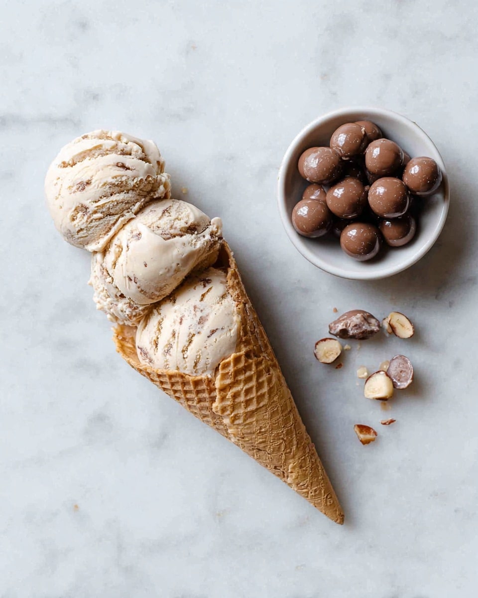 A three-scoop ice cream cone lies flat on a white marbled surface, with the cone on the left and melting swirled beige and brown ice cream scoops stacked closely together, showing a creamy and slightly textured appearance. To the top right, there is a small white bowl filled with smooth, round chocolate-covered malt balls, some of which are broken to reveal the light center inside. A few broken pieces of these malt balls are scattered near the bowl on the surface. photo taken with an iphone --ar 4:5 --v 7
