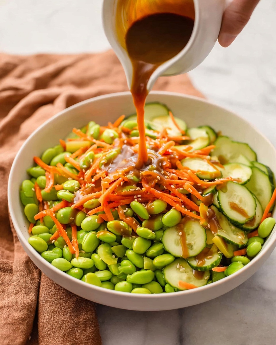 A white bowl is filled with a fresh salad that has three main layers: bright green edamame beans scattered all over the bowl, thin orange carrot sticks mixed throughout, and light green cucumber slices with darker green edges spread on top. A woman's hand is pouring a rich brown dressing over the salad from a white small pitcher, the dressing flowing in a smooth stream onto the vegetables. The bowl sits on a white marbled surface with a soft brown cloth nearby. photo taken with an iphone --ar 4:5 --v 7