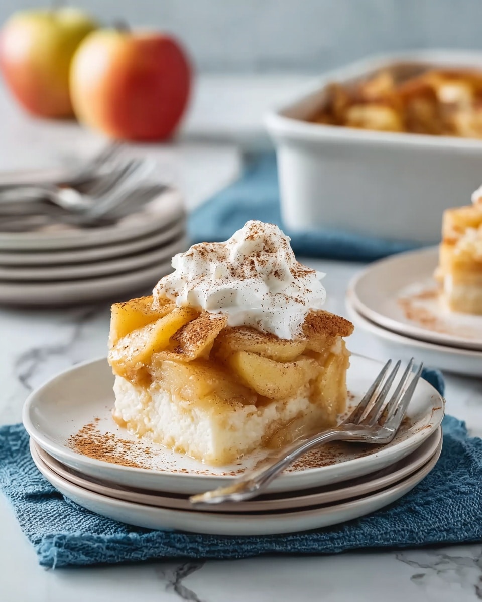 A square piece of apple dessert with three visible layers sits on a stack of three white plates. The bottom layer is a soft, pale cake base. The middle layer consists of cooked apple slices in a light syrupy sauce, showing a golden-yellow and slightly translucent color. The top layer has more apple slices, browned and sprinkled with cinnamon powder, and a dollop of white whipped cream slightly melting, also dusted with cinnamon on top. A fork rests on the right side of the plate. This scene is set on a white marbled surface with a blue cloth partially visible underneath the plates. In the blurred background, there is a white baking dish filled with more dessert and two apples, with a stack of white plates with silver forks beside it. Photo taken with an iphone --ar 4:5 --v 7