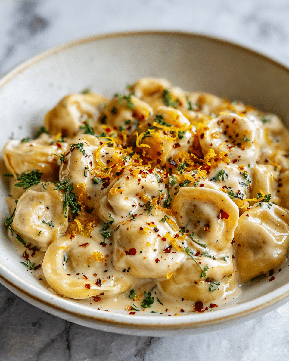 The image shows a close-up of small round pasta shapes, possibly tortellini, covered in a creamy beige sauce. The pasta pieces have a smooth, slightly shiny texture and are nestled closely together in a white bowl. Scattered on top are small bits of bright yellow zest and red pepper flakes that add color contrast and texture to the dish. The creamy sauce looks thick and coats each pasta piece evenly, with a few tiny black specks visible within it. The setting includes a white marbled surface in the background, adding a clean and elegant feel. photo taken with an iphone --ar 4:5 --v 7