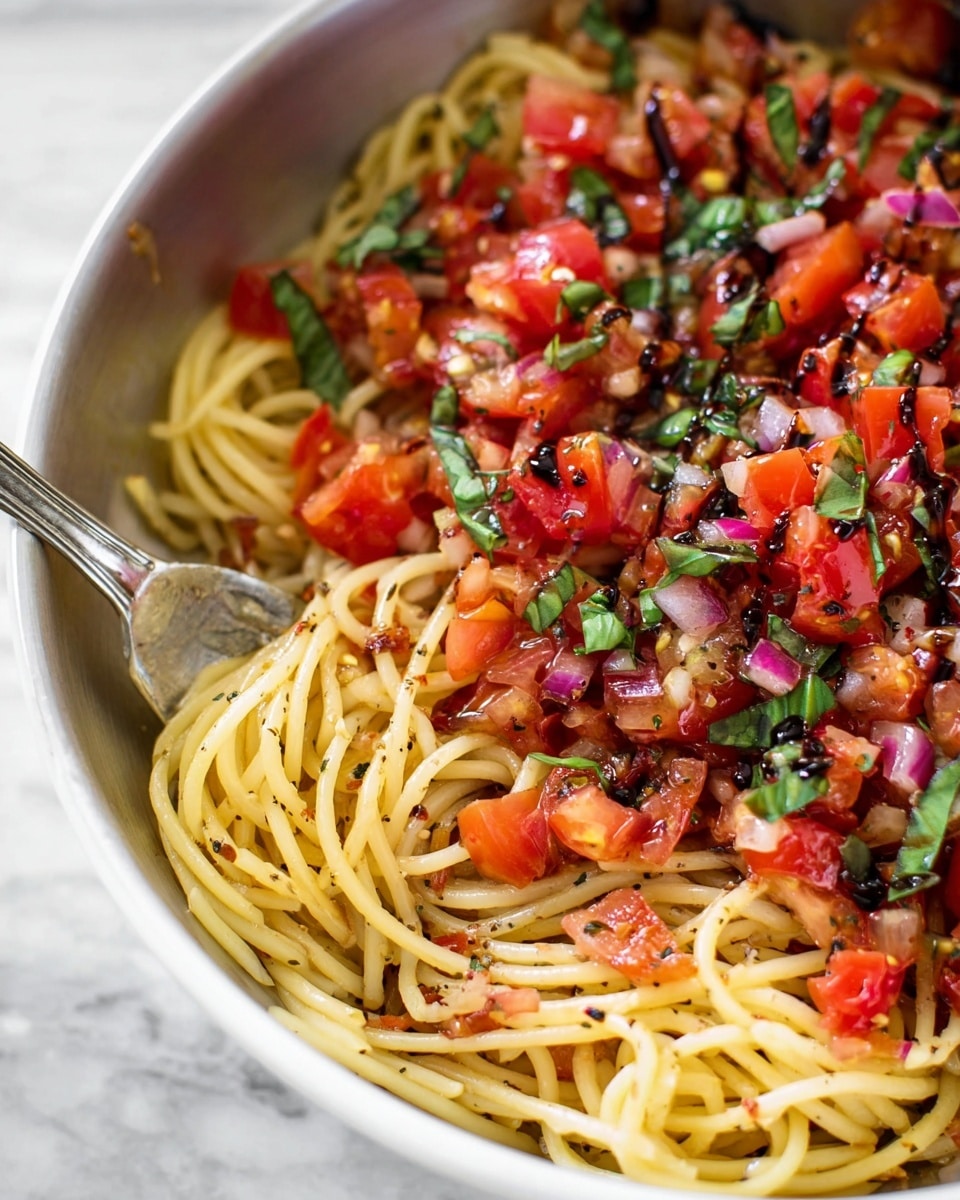 A close-up showing a white bowl filled with spaghetti pasta that is lightly coated in oil and speckled with black pepper, forming a yellow twisted base layer. On top, there is a thick layer of diced bright red tomatoes mixed with finely chopped purple onions and dark green basil leaves scattered throughout. The tomatoes and onions have a glossy look, and the whole dish is drizzled with a dark balsamic glaze that adds a shiny contrast to the fresh ingredients. A metal fork is inserted into the spaghetti on the left side, stirring the pasta slightly. The bowl sits on a white marbled surface. Photo taken with an iphone --ar 4:5 --v 7