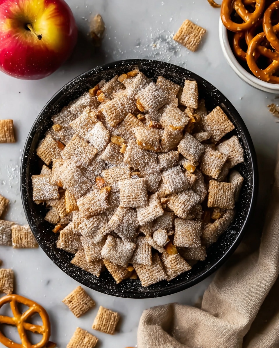A black bowl filled with many small square cereal pieces, light brown in color with a rough texture, and small nut-like bits embedded on some pieces, all lightly dusted with white powdered sugar. The bowl sits on a white marbled surface, with some powdered sugar scattered around it and a few cereal pieces outside the bowl. To the left, a red and yellow apple is partially visible, and to the right, part of a white bowl filled with pretzels can be seen. A beige cloth is draped slightly on the upper right side of the image. Photo taken with an iphone --ar 4:5 --v 7
