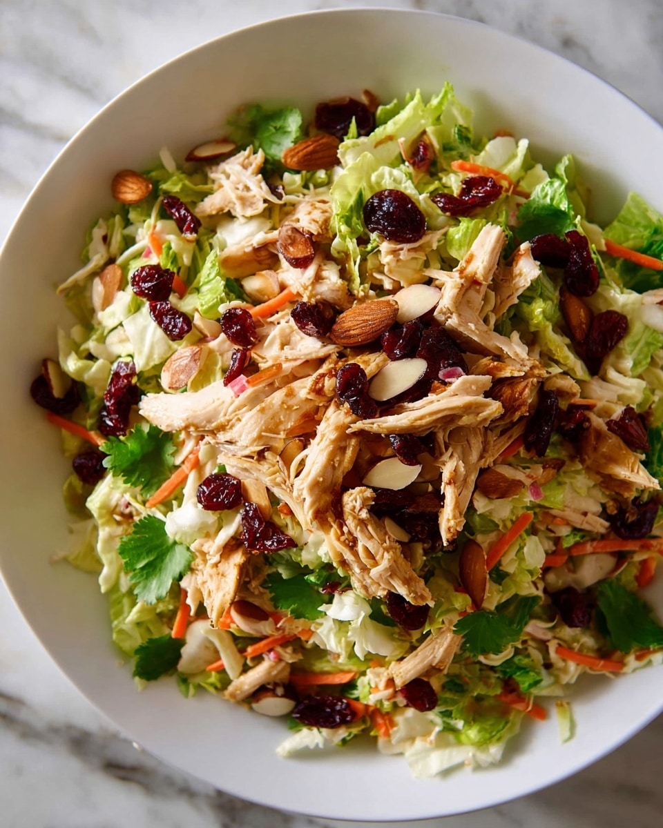 The image shows a fresh salad in a white bowl on a white marbled texture. The salad has a base layer of light and dark green leafy lettuce mixed with shredded white cabbage and thin orange carrot strips spread evenly. On top of this, there are pieces of shredded cooked chicken with a light brown color. Scattered over the top are dark red dried cranberries, sliced light brown almonds, and a few whole almonds, adding texture and color contrast. Some small green cilantro leaves are spread across, adding freshness. The salad looks vibrant and crunchy with varied textures and colors mixing green, orange, red, and brown tones. photo taken with an iphone --ar 4:5 --v 7