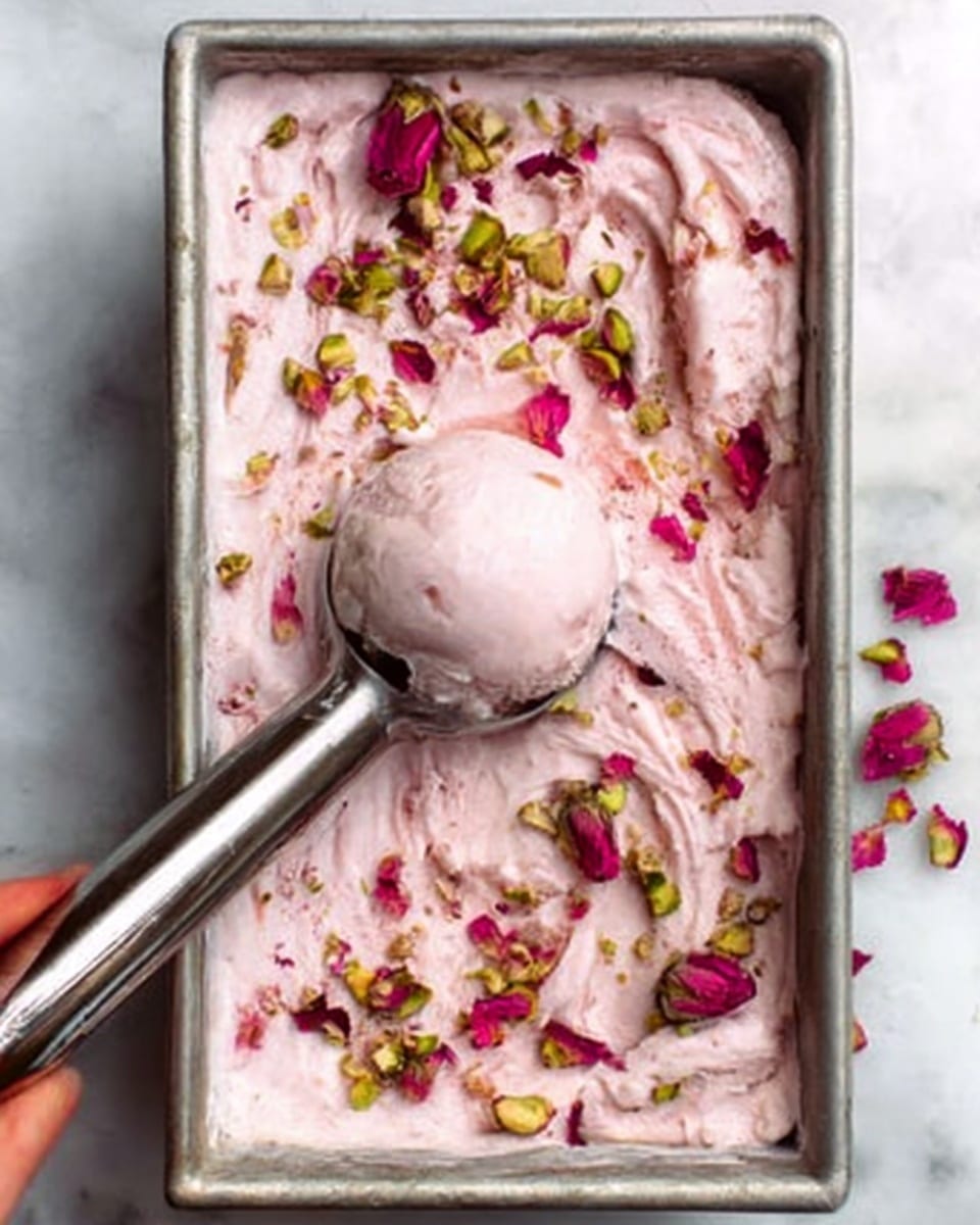 The image shows a rectangular metal container filled with creamy pink rose-flavored ice cream topped with scattered rose petals and sprinkled with crushed pistachios. The ice cream has visible swirls and soft texture throughout the top layer. A scoop of ice cream is lifted in a shiny metal ice cream scooper, revealing smooth and creamy inside. A woman's hand holds the scooper, gently lifting it from the container. The scene is set on a white marbled surface. Photo taken with an iphone --ar 4:5 --v 7