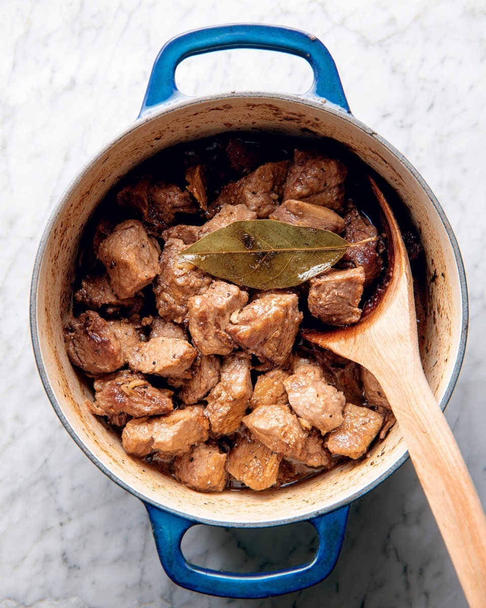A blue pot filled with cooked brown meat chunks that have a slightly crispy texture on the outside, mixed evenly inside a light cream interior pot. The meat pieces vary in size and are stacked loosely with a visible single brown bay leaf placed on top near the center. A wooden spoon with a smooth, rounded end rests inside the pot on the right side, partially touching the meat. The pot sits on a white marbled surface showing subtle gray veins with a soft, natural light falling from above. photo taken with an iphone --ar 4:5 --v 7