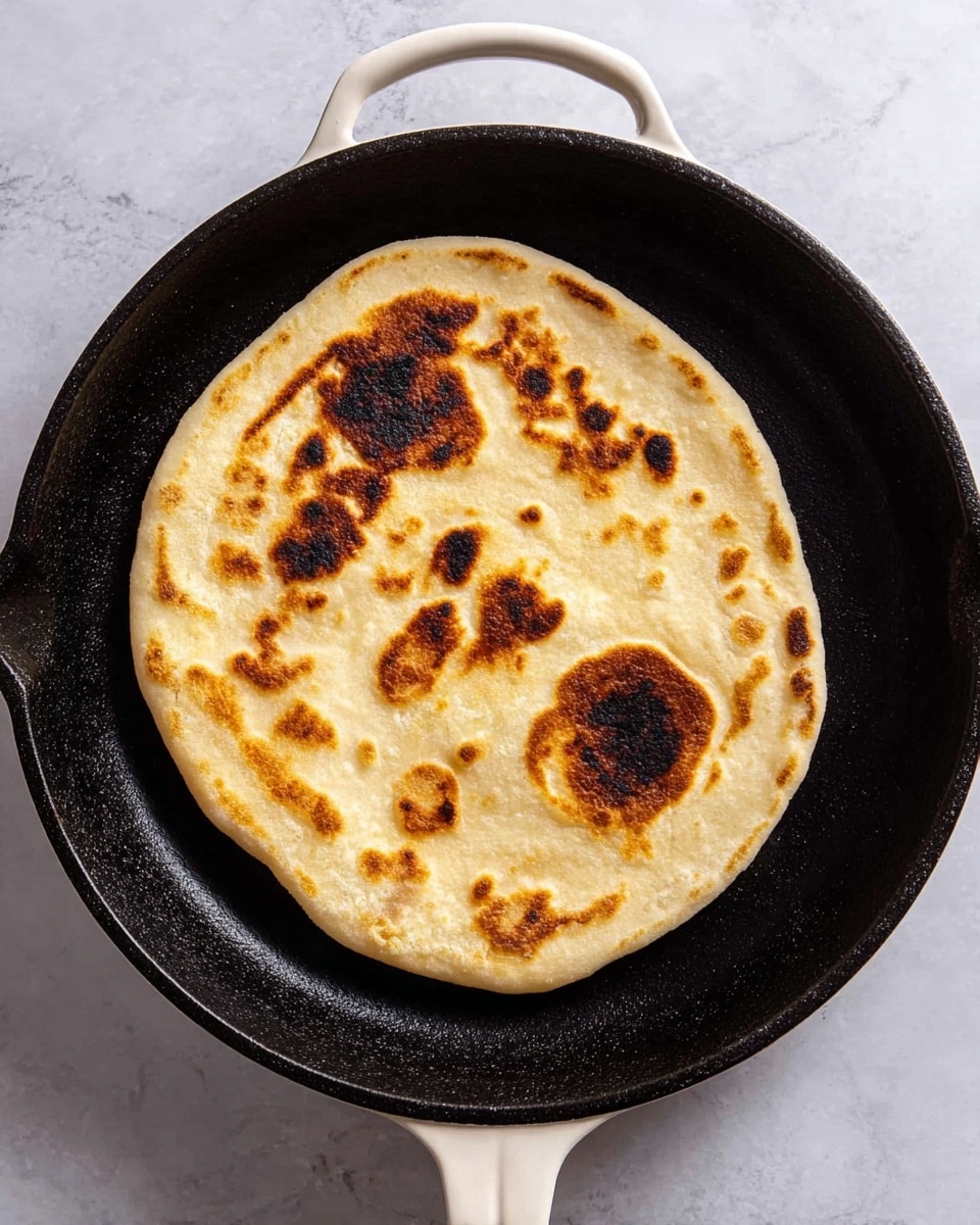 A round flatbread with an uneven shape is cooking in a black cast iron skillet with a white handle. The flatbread has a golden brown surface with darker brown spots, especially two large, distinct marks near one edge, showcasing a toasted texture. The bread appears soft and thick, slightly puffed up in some areas. The skillet rests on a white marbled surface. photo taken with an iphone --ar 4:5 --v 7