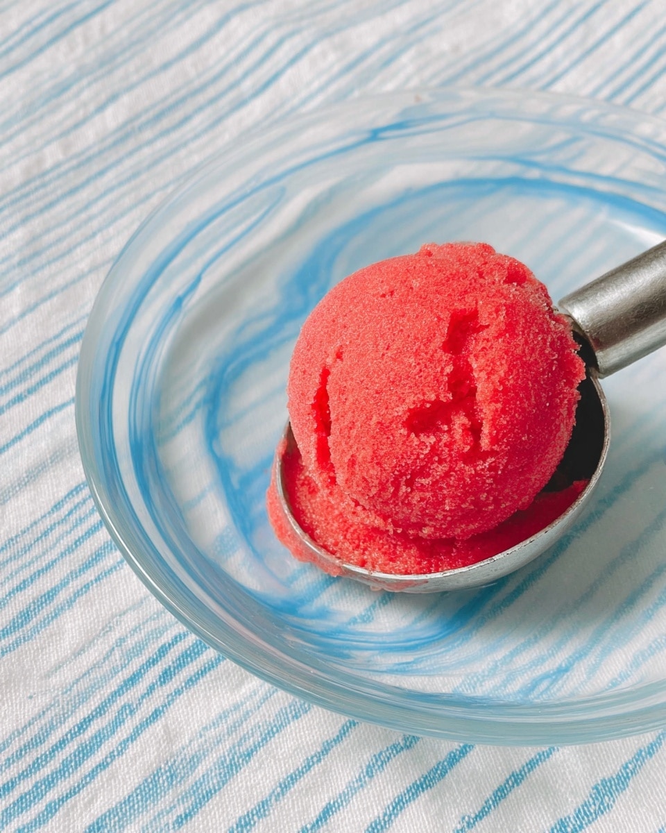 A close-up view of a single scoop of bright red sorbet with a slightly rough texture, placed on a shiny metal ice cream scoop. The sorbet is round and looks cold and fresh. The scoop rests on a clear, white plate with blue swirl patterns, sitting on a white marbled texture with light blue and white striped cloth underneath. photo taken with an iphone --ar 4:5 --v 7