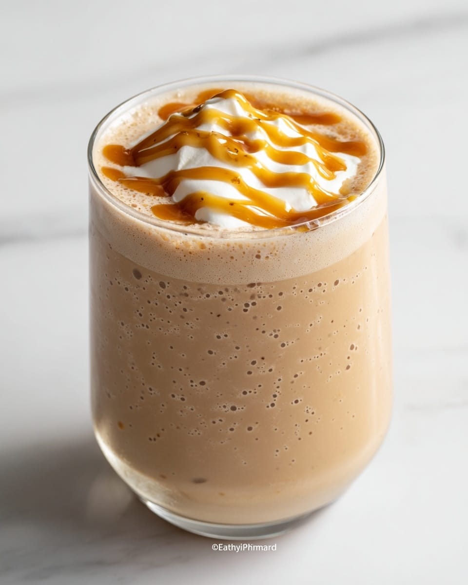 A creamy light brown drink fills a clear glass almost to the top, with small bubbles throughout the texture. On the surface, there is a dollop of white whipped cream that is swirled with rich golden caramel sauce in a zigzag pattern. The glass is set on a white marbled surface, showing smooth, soft textures in the drink and the shiny, smooth glass edges. photo taken with an iphone --ar 4:5 --v 7
