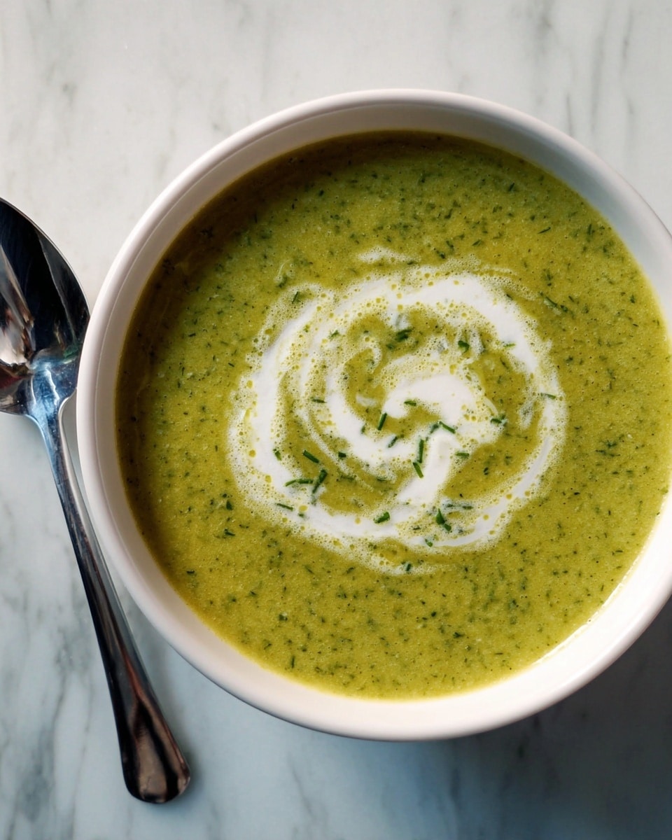 A white bowl filled with smooth, green soup that has small bits of herbs visible throughout. On the top surface of the soup, there is a swirl of white cream forming a loose spiral pattern near the center. The bowl sits on a white marbled surface. A shiny metal spoon is placed to the left side of the bowl. photo taken with an iphone --ar 4:5 --v 7