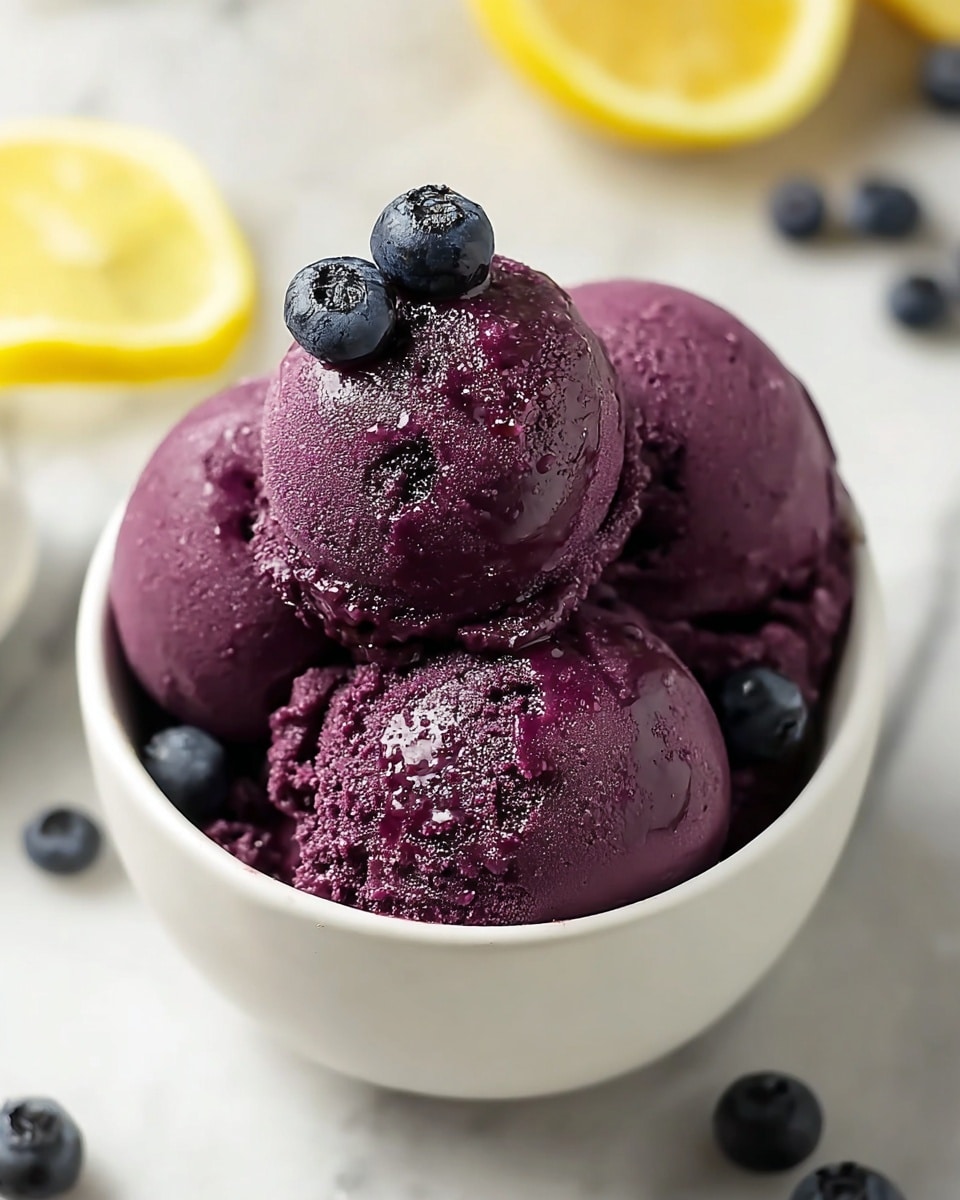 A white bowl filled with three smooth scoops of dark purple ice cream with a slightly shiny, creamy texture, topped with three fresh blueberries resting on the top scoop. The ice cream surface shows subtle bits of berry and a few moisture droplets, giving it a fresh look. The bowl is placed on a white marbled surface with a few scattered blueberries around it and blurred lemon slices in the background. Photo taken with an iphone --ar 4:5 --v 7