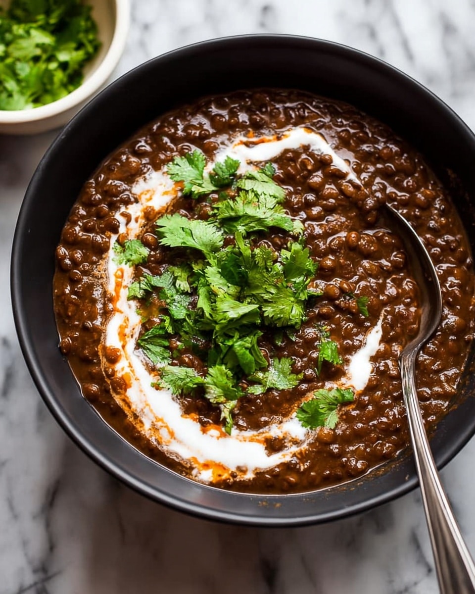A close-up view of a black bowl filled with a thick, dark brown lentil stew that has a slightly chunky texture. On top, there is a swirl of white cream unevenly spread across the surface, adding a smooth and light contrast. Fresh green cilantro leaves cover part of the cream, placed mainly in the middle, providing a bright, leafy accent against the dark stew. A silver spoon is partially submerged on the right side of the bowl, reflecting light and resting against the bowl's edge. The bowl is placed on a white marbled surface with a small white bowl of cilantro leaves blurred in the background. photo taken with an iphone --ar 4:5 --v 7