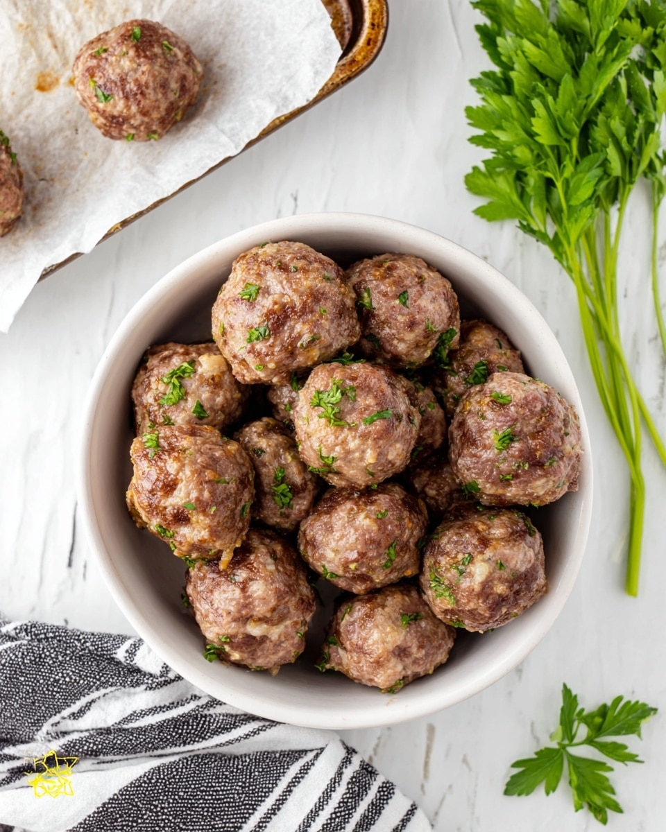 A white bowl filled with around fifteen round meatballs, each showing a browned, slightly textured surface with small bits of onion visible. The meatballs are garnished lightly with chopped green parsley scattered over them. Part of a baking tray with two more meatballs on white parchment paper is visible above the bowl, showing a slightly greasy texture on the paper. Fresh green parsley stems lie to the right of the bowl on a white marbled surface. A white and dark striped cloth is partly visible under the bowl at the bottom left corner. Photo taken with an iphone --ar 4:5 --v 7
