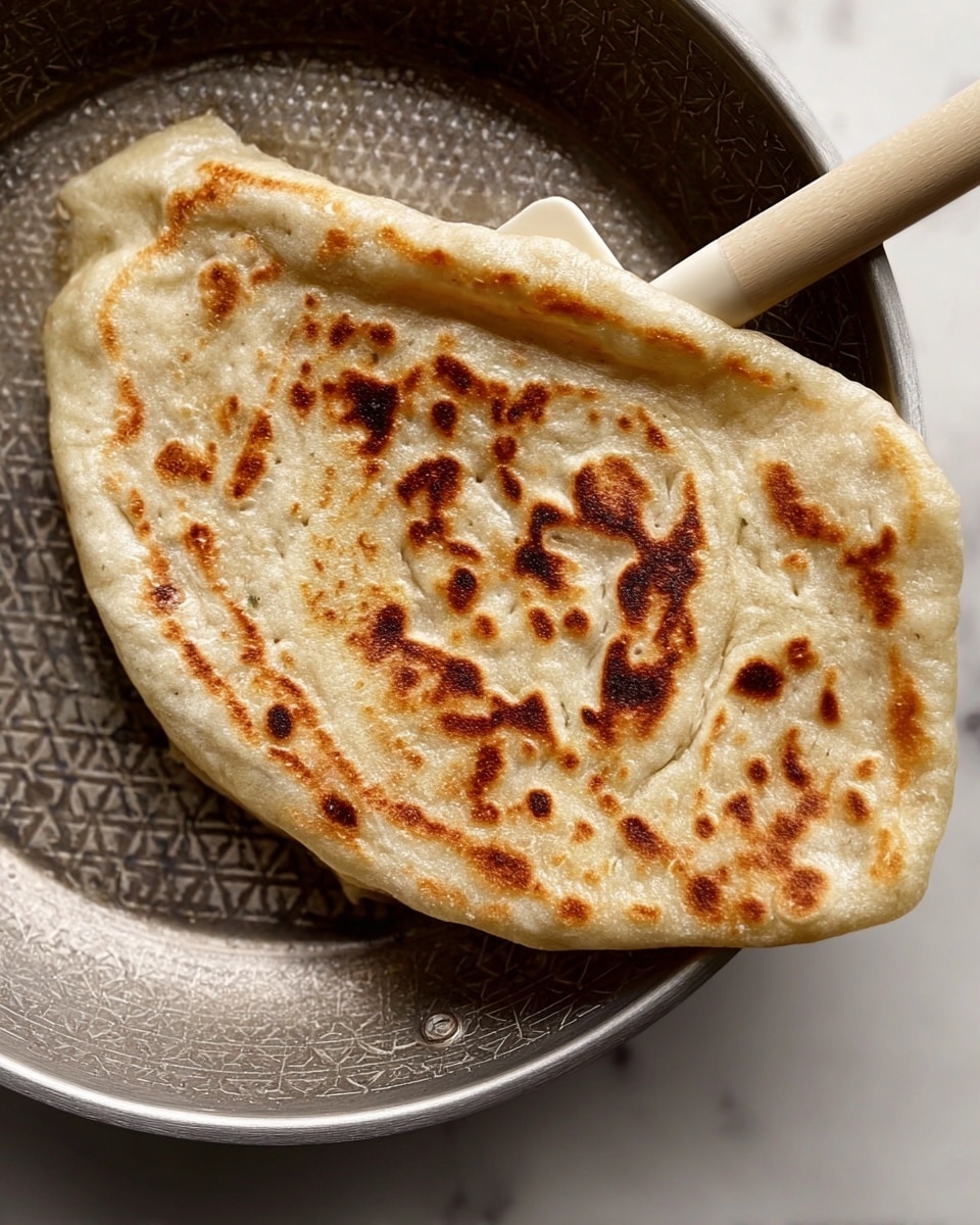 A close-up of a folded flatbread cooked with a golden-brown, slightly uneven toasted pattern on its surface, showing darker brown spots scattered mostly in the center and edges. The flatbread has a soft, slightly bubbled texture. It is held by a beige spatula resting inside a metal frying pan with a hexagonal patterned center, placed on a white marbled texture surface. The angle shows the thick edges and soft inside of the flatbread with some air bubbles beneath it. photo taken with an iphone --ar 4:5 --v 7