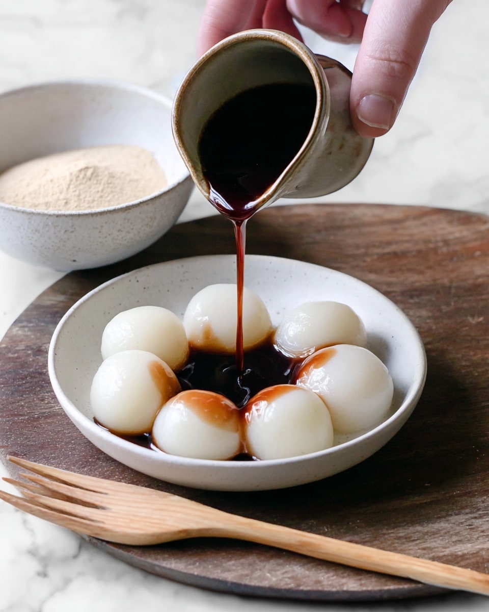 A small white bowl holds seven smooth, round, glossy white mochi balls neatly arranged in a single layer. A woman's hand pours a thick dark brown syrup from a small ceramic cup over the mochi, the syrup dripping in a thin stream creating shiny dark patches on the white mochi surface. In the background, a white bowl with light tan powder is visible on a white marbled surface, and a light wooden fork lies in front of the bowl. photo taken with an iphone --ar 4:5 --v 7