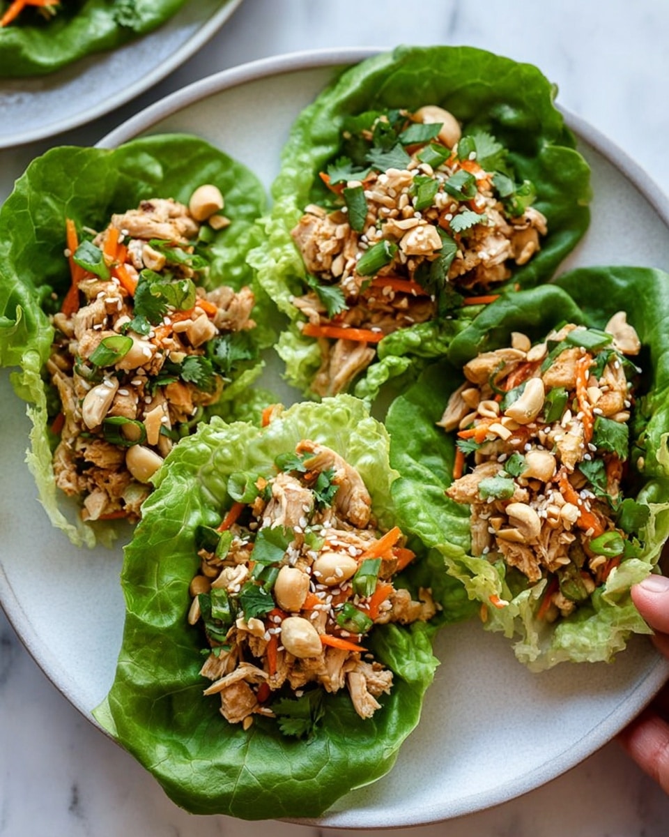 Three lettuce wraps sit on a white plate with a white marbled background. Each wrap uses a bright green lettuce leaf as the base, fresh and textured with ruffled edges. Inside, shredded cooked chicken mixed with small orange carrot pieces, chopped green scallions, and fresh cilantro leaves form a colorful, chunky layer. The chicken mixture is topped with scattered light beige chopped cashews and white sesame seeds, adding a crunchy texture. A woman's hand is gently holding one of the lettuce wraps on the right edge of the plate. Photo taken with an iphone --ar 4:5 --v 7
