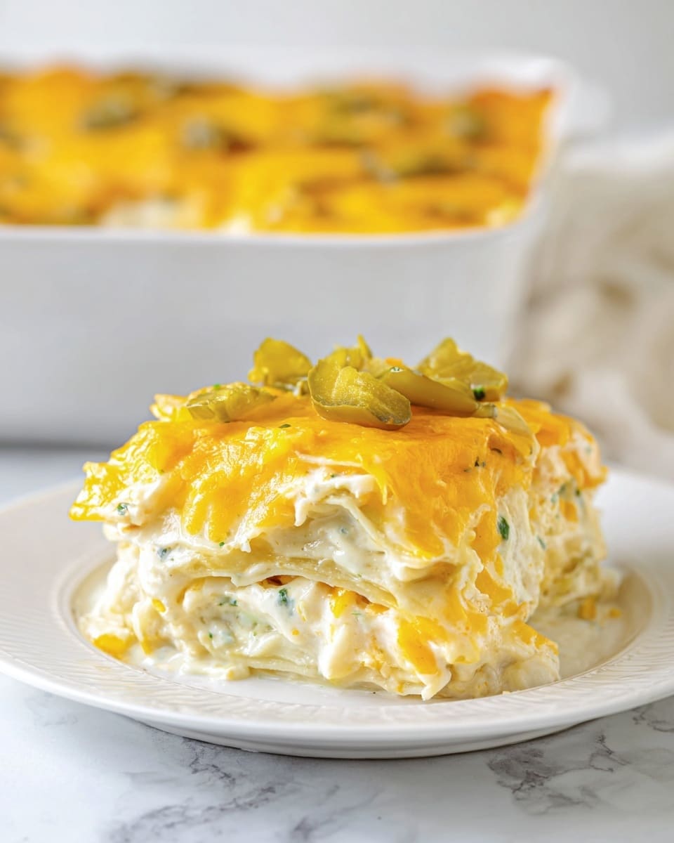 The image shows a thick slice of creamy layered casserole on a white plate placed on a white marbled surface. The bottom layer is pale yellow pasta or tortilla, followed by a thick layer of white creamy sauce mixed with green herbs. Above this is another pasta or tortilla layer covered generously with melted orange cheddar cheese pieces and small bits of green pickles on top. The dish looks soft, rich, and cheesy with a slightly textured surface from the melted cheese and pickles. In the background, a white baking dish filled with more of the same casserole is slightly out of focus. photo taken with an iphone --ar 4:5 --v 7